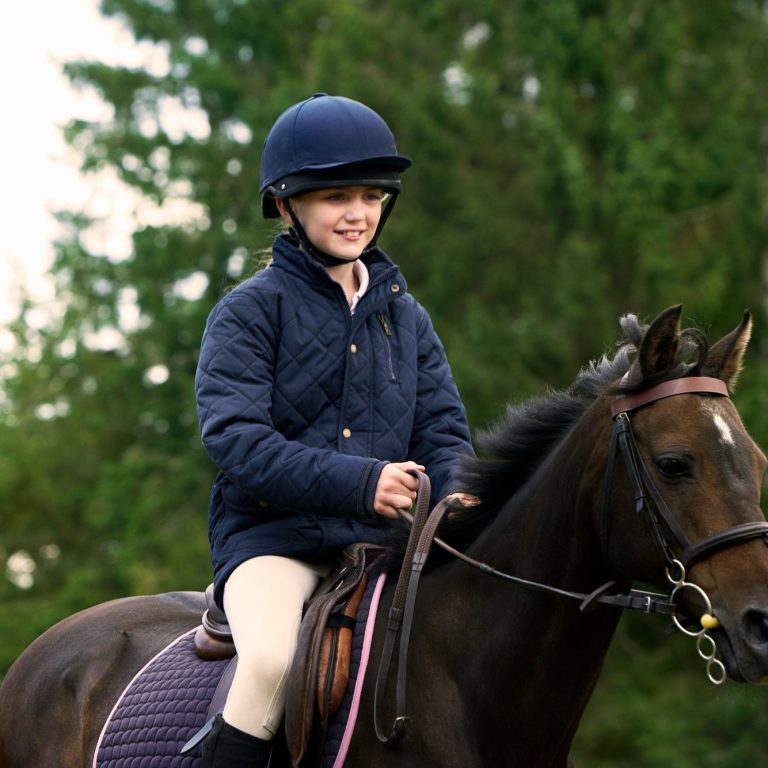 Ein lächelndes Kind auf einem Pferd, trägt einen Helm und eine Reitjacke.