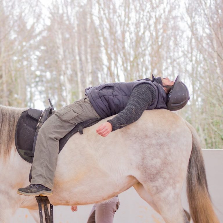 Person liegt entspannt auf einem Pferd in einer Reithalle.